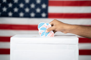 Close up of Hands dropping multiple I Voted sticker inside Ballot box with US flag as background, Concept of fraud in USA elections